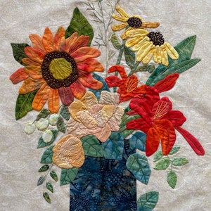 May include: A fabric art piece featuring a vibrant floral arrangement in a blue vase. The bouquet includes a large sunflower, yellow coneflowers, and red lilies, with green leaves and stems. The background is a light beige fabric with a subtle pattern.
