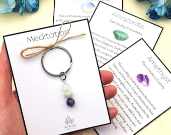 Puede incluir: Un llavero de plata con tres cuentas de cristal: una ópalo azul claro, una aventurina verde y una amatista púrpura. El llavero está en una tarjeta blanca con la palabra "Meditación" y el texto "KORONI CREATIONS".