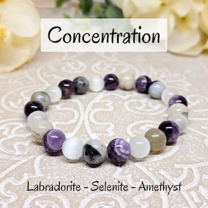 Op de afbeelding: Een armband gemaakt met Labradorite, Seleniet en Amethist kralen. De armband is wit, grijs en paars. De tekst "Concentration" staat boven de armband en de tekst "Labradorite - Selenite - Amethyst" staat onder de armband.