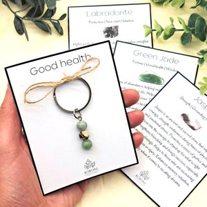 May include: A keychain with three green and brown gemstone beads attached to a silver ring. The keychain is on a white card with the text "Good health" and the name of the company "KORONI CREATIONS".