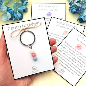 Puede incluir: Un llavero con tres cuentas de cristal en tonos blanco, rosa y azul. Las cuentas están unidas a un anillo de llave plateado con un lazo de cuerda marrón. El llavero se muestra en una tarjeta blanca con el texto "Peace and Calm" y el logotipo "KORONI CREATIONS".