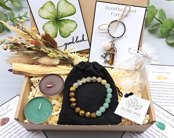 May include: A gift box filled with items for good luck and fortune. The box contains a green four-leaf clover card, a sage smudge stick, two tealight candles, a green and brown beaded bracelet, a clear crystal, a keychain with a crystal pendant, and several cards with text about the crystals. The text on the cards reads "Good Luck and Fortune", "Citrine", and "Green Aventurine".