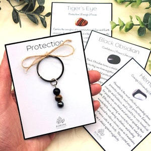 Puede incluir: Un llavero de plata con tres cuentas: negra, ojo de tigre y hematites. El llavero está en una tarjeta blanca con el texto "Protección" en letras negras. La tarjeta también tiene el texto "Koroni Creations" en letras negras.