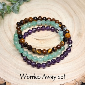 May include: Three stacked bracelets made of amethyst, green aventurine, and tiger's eye beads. The bracelets are on a wooden surface and the text "Worries Away set" is below the bracelets.