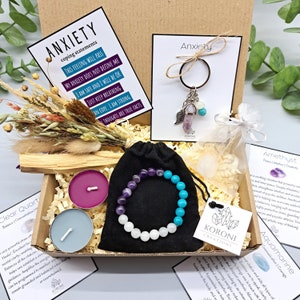 Peut inclure: Un coffret cadeau pour le soulagement de l'anxiété. Il comprend un bracelet avec des perles d'améthyste et de turquoise, deux bougies, un porte-clés avec des breloques, des fleurs séchées et des cartes d'affirmation. La boîte est remplie de copeaux de bois.
