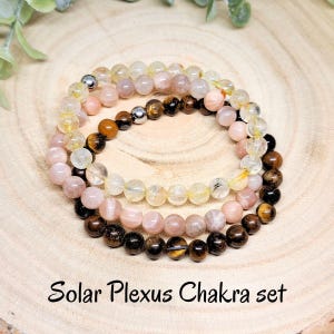 Può includere: Tre braccialetti di perline in un set. I braccialetti sono fatti di perline rosa chiaro, trasparenti e marroni. Il testo "Solar Plexus Chakra set" è scritto sotto i braccialetti.