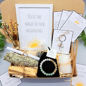 May include: A gift box filled with spiritual items. It includes a framed print with the text "Trust the Magic of New Beginnings", a sage bundle, palo santo sticks, a bracelet, a candle, and a keychain.