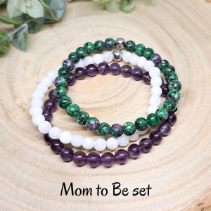 Könnte beinhalten: Drei Armbänder, die auf einer Holzoberfläche gestapelt sind. Das obere Armband ist grün mit rosa Flecken, das mittlere Armband ist weiß und das untere Armband ist lila. Der Text "Mom to Be set" steht unter den Armbändern.