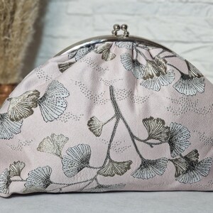 Könnte beinhalten: Hellrosa Clutch mit silbernem Verschluss. Sie zeigt ein sich wiederholendes Muster aus silbernen und hellblauen Ginkgoblättern auf dunkelgrauen Stielen. Das Design bildet ein elegantes Blumenmuster.
