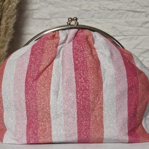 Könnte beinhalten: Eine Clutch mit silbernem Verschluss und vertikalem Streifenmuster in Rosa-, Weiß- und Koralltönen. Der Stoff hat einen dezenten Schimmer. Die Tasche hat eine abgerundete Form.