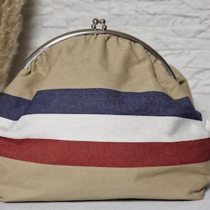 Könnte beinhalten: Beige Stoff-Clutch mit silbernem Verschluss. Die Clutch hat horizontale Streifen in Marineblau, Weiß und Rot. Die Tasche hat eine abgerundete Form und scheint aus einem strukturierten Material zu bestehen.
