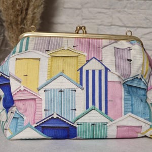 Könnte beinhalten: Stoff-Clutch mit einem bunten Strandhütten-Design. Die Hütten sind in Pastelltönen wie Gelb, Rosa, Blau und Grün gehalten, mit weißen Details. Sie hat einen goldfarbenen Verschluss und Rahmen.