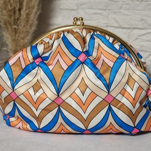 Könnte beinhalten: Eine farbenfrohe Clutch mit goldenem Verschluss. Der Stoff zeigt ein geometrisches Muster in Blau-, Orange-, Rosa-, Braun- und Weißtönen. Die Clutch hat eine abgerundete Form.