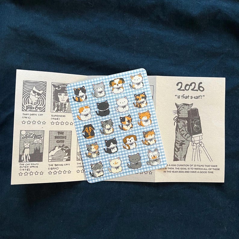 Puede incluir: Un conjunto de art&iacute;culos con tem&aacute;tica de gatos. Una hoja de pegatinas con ilustraciones de gatos sobre un fondo de cuadros azules y dos tarjetas plegadas. Una tarjeta presenta t&iacute;tulos de pel&iacute;culas sobre gatos, la otra dice "2026 is that a cat?"