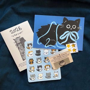 May include: A collection of cat-themed stationery and stickers. Includes a blue card with a black cat illustration and a light blue bow, a small notepad with the text "2026", and a sheet of cat stickers on a blue gingham background.