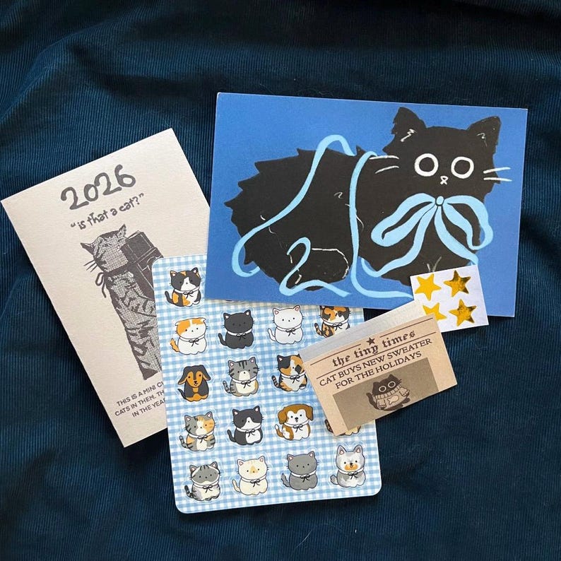 Puede incluir: Una colecci&oacute;n de art&iacute;culos de papeler&iacute;a y pegatinas con tem&aacute;tica de gatos. Incluye una tarjeta azul con una ilustraci&oacute;n de gato negro y un lazo azul claro, un peque&ntilde;o bloc de notas con el texto "2026" y una hoja de pegatinas de gatos.
