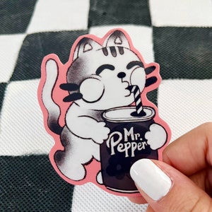 May include: A pink and white cartoon cat sticker with black outlines, holding a can of Mr. Pepper with a straw in its mouth. The sticker is on a black and white checkered background.