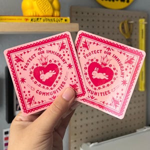 May include: Two pink square stickers with a decorative border. The stickers feature a heart with a rabbit inside, surrounded by the text "PROTECT OUR IMMIGRANT COMMUNITIES". Additional floral and star designs are included.