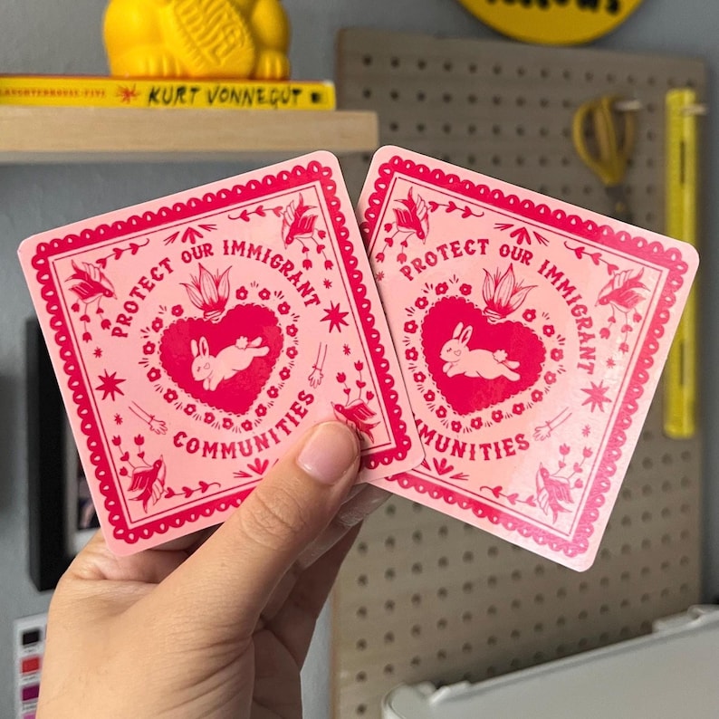 May include: Two pink square stickers with a decorative border. The stickers feature a heart with a rabbit inside, surrounded by the text "PROTECT OUR IMMIGRANT COMMUNITIES". Additional floral and star designs are included.