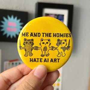 May include: A yellow button with black text and illustrations. The text reads "ME AND THE HOMIES HATE AI ART." Three cartoon cat figures hold hands and knives. The button is round and held by a hand.