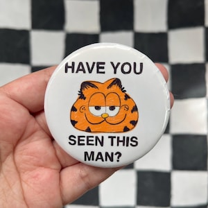 May include: A white button featuring the cartoon character Garfield with the text "HAVE YOU SEEN THIS MAN?". Garfield is orange with black stripes and a smiling expression. The button is held against a black and white checkered background.