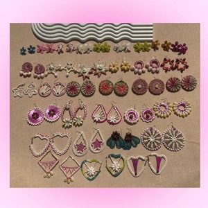 May include: A collection of handmade earrings in various shapes and colors, including floral, star, and geometric designs. The earrings feature intricate woven and beaded details, with a range of pink, white, and green hues. The earrings are displayed on a neutral surface.