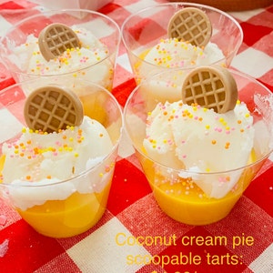 Pu&ograve; includere: Quattro bicchieri trasparenti pieni di crema pasticcera gialla, sormontati da panna montata bianca, zuccherini colorati e una decorazione rotonda a forma di waffle marrone. I bicchieri sono su una superficie a scacchi rossi e bianchi. Testo: Crostate di crema di cocco.