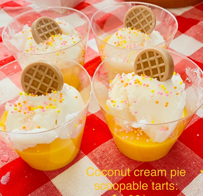 Pu&ograve; includere: Quattro bicchieri trasparenti pieni di crema pasticcera gialla, sormontati da panna montata bianca, zuccherini colorati e una decorazione rotonda a forma di waffle marrone. I bicchieri sono su una superficie a scacchi rossi e bianchi. Testo: Crostate di crema di cocco.