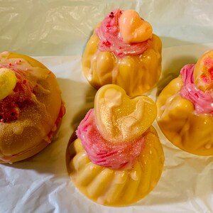 Pu&ograve; includere: Quattro saponi decorativi a forma di cupcake con glassa rosa e decorazioni a forma di cuore. I saponi sono marrone chiaro con una lucentezza dorata e la glassa &egrave; cosparsa di zuccherini. I saponi sono disposti su una superficie bianca.