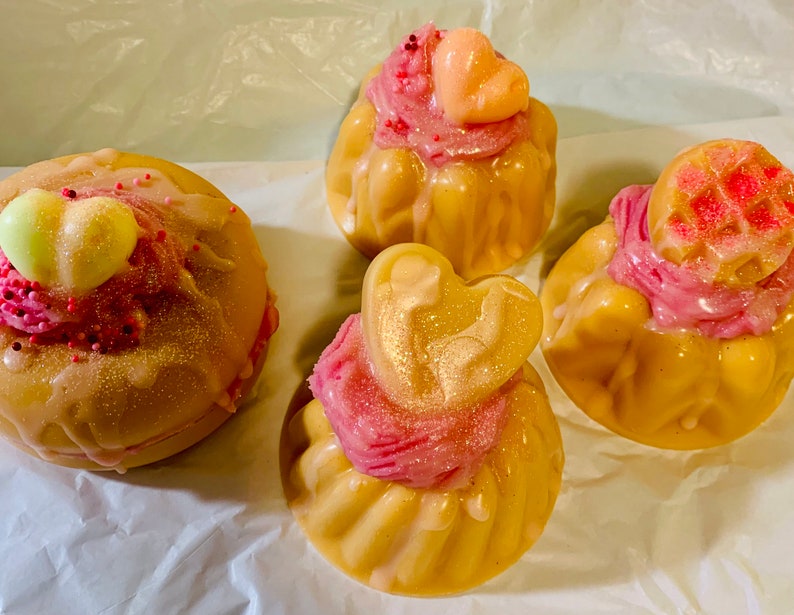 Pu&ograve; includere: Quattro saponi decorativi a forma di cupcake con glassa rosa e decorazioni a forma di cuore. I saponi sono marrone chiaro con una lucentezza dorata e la glassa &egrave; cosparsa di zuccherini. I saponi sono disposti su una superficie bianca.