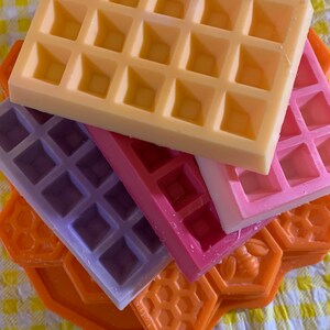 Pu&ograve; includere: Una pila di cera profumata colorata a forma di waffle e favi. I waffle sono gialli, rosa, viola e rossi. I favi sono arancioni. Lo sfondo &egrave; un motivo a scacchi giallo e bianco.