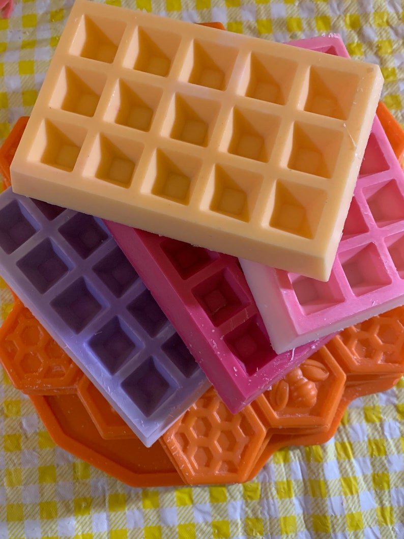 Pu&ograve; includere: Una pila di cera profumata colorata a forma di waffle e favi. I waffle sono gialli, rosa, viola e rossi. I favi sono arancioni. Lo sfondo &egrave; un motivo a scacchi giallo e bianco.