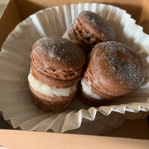 Pu&ograve; includere: Primo piano di macarons al cioccolato con ripieno bianco, cosparsi di glitter, all'interno di una scatola di cartone marrone. I macarons sono disposti su un rivestimento di carta bianca. L'immagine evidenzia la consistenza e il colore del dessert.