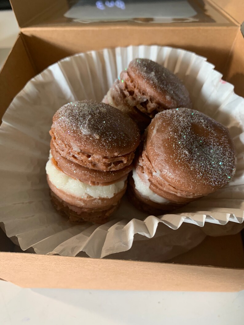 Pu&ograve; includere: Primo piano di macarons al cioccolato con ripieno bianco, cosparsi di glitter, all'interno di una scatola di cartone marrone. I macarons sono disposti su un rivestimento di carta bianca. L'immagine evidenzia la consistenza e il colore del dessert.