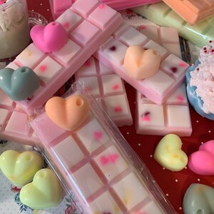 Pu&ograve; includere: Una collezione di cere profumate e cere a forma di cuore. Le cere sono in vari colori, tra cui rosa, pesca e verde. Alcune cere sono decorate con piccole forme di cuore. Le cere sono disposte su una superficie a pois rossi e bianchi.