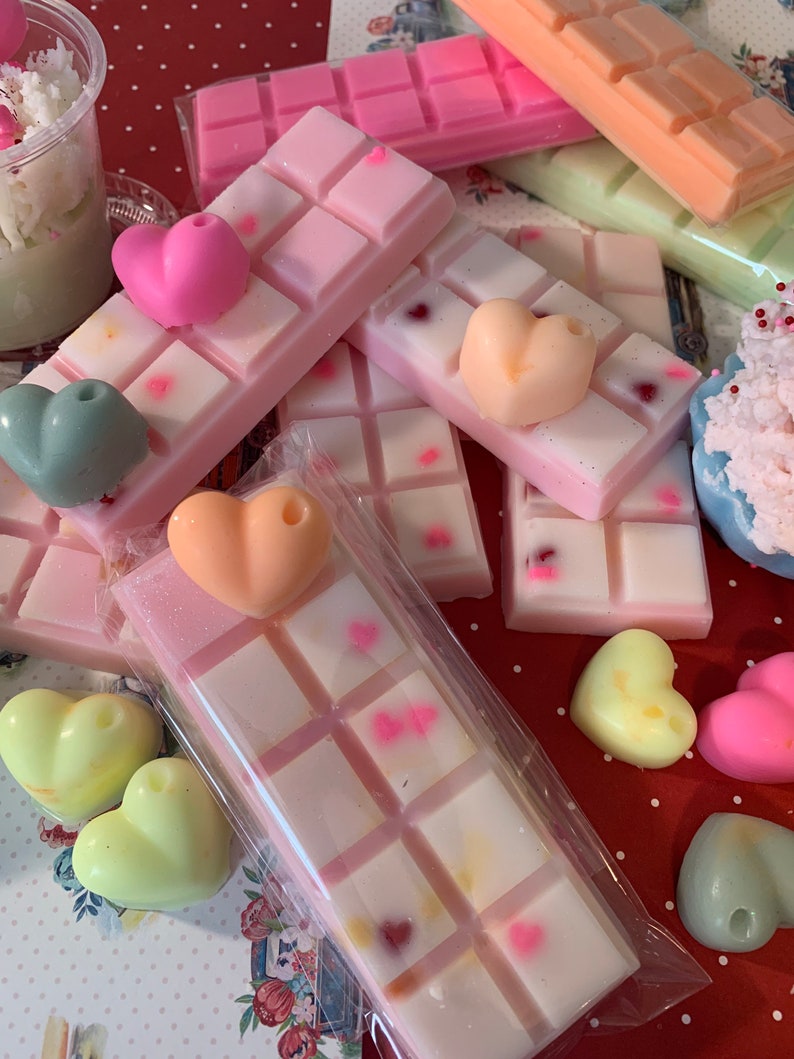 Pu&ograve; includere: Una collezione di cere profumate e cere a forma di cuore. Le cere sono in vari colori, tra cui rosa, pesca e verde. Alcune cere sono decorate con piccole forme di cuore. Le cere sono disposte su una superficie a pois rossi e bianchi.