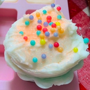 Pu&ograve; includere: Un sapone a forma di cupcake bianco, con una leggera spolverata arancione e spruzzi colorati. Il sapone &egrave; in uno stampo rosa, con un motivo a scacchi rossi e bianchi sullo sfondo. Il sapone ha un aspetto strutturato e smerigliato.