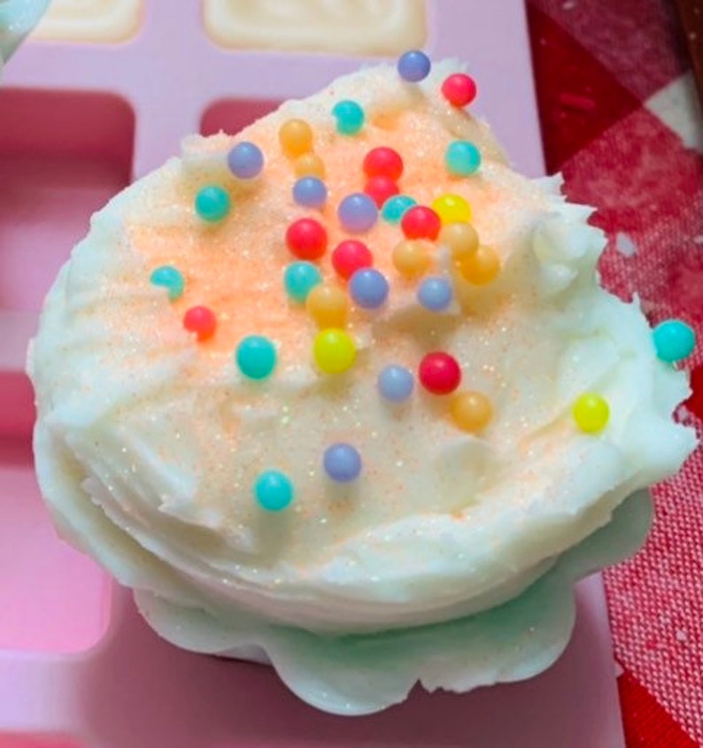 Pu&ograve; includere: Un sapone a forma di cupcake bianco, con una leggera spolverata arancione e spruzzi colorati. Il sapone &egrave; in uno stampo rosa, con un motivo a scacchi rossi e bianchi sullo sfondo. Il sapone ha un aspetto strutturato e smerigliato.