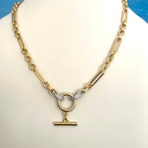 May include: A gold chain necklace with a toggle clasp. The chain is made of two different styles of links, one is a round link and the other is a rectangular link. The links are accented with small clear stones.