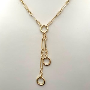 May include: A gold-colored necklace with a Y-shaped design. The necklace features a series of elongated links and two dangling chains with circular and rectangular elements. The necklace is displayed on a white surface.