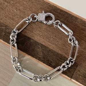 May include: A silver-toned bracelet with a rectangular link chain design. The clasp is encrusted with small, clear stones. The bracelet is displayed on a wooden surface.