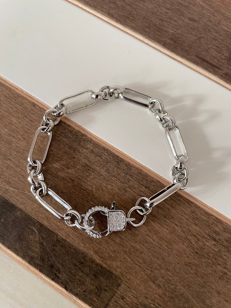 May include: Silver-toned bracelet featuring a chain link design with a rectangular clasp. The clasp is adorned with small, sparkling stones. The bracelet is displayed on a wooden surface with white accents.