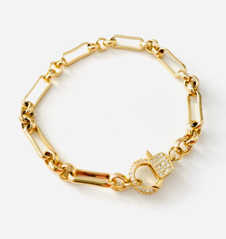 May include: Gold-toned bracelet featuring a mix of chain links and rectangular bars. The bracelet has a decorative clasp encrusted with small, clear crystals. A stylish piece of jewelry.