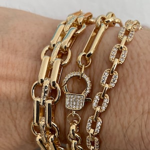 May include: Gold-toned chain bracelets in various designs. One bracelet features a series of rectangular links, another has a padlock clasp, and the third is adorned with small, sparkling stones. The bracelets are displayed on a wrist.