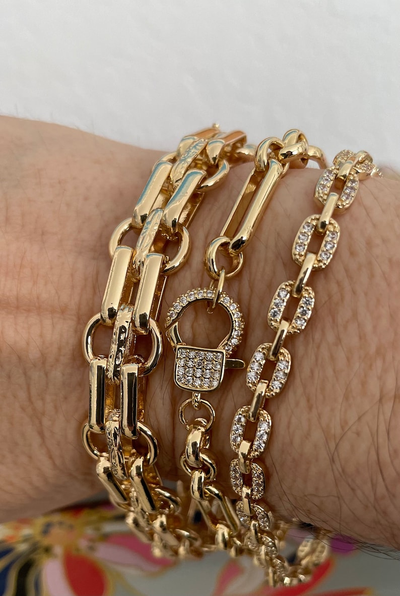 May include: Gold-toned chain bracelets in various designs. One bracelet features a series of rectangular links, another has a padlock clasp, and the third is adorned with small, sparkling stones. The bracelets are displayed on a wrist.