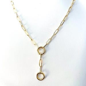 May include: A gold-colored necklace with a Y-shaped design. The necklace features a chain with elongated oval links. A large ring connects the chain to a shorter chain with a small ring at the end. The necklace is displayed against a white background.
