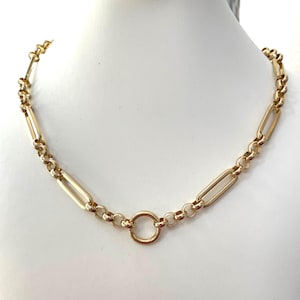 May include: A gold-toned chain necklace featuring a repeating pattern of elongated rectangular links alternating with smaller circular links. The necklace is centered with a single, prominent circular ring connector that joins the two sides.