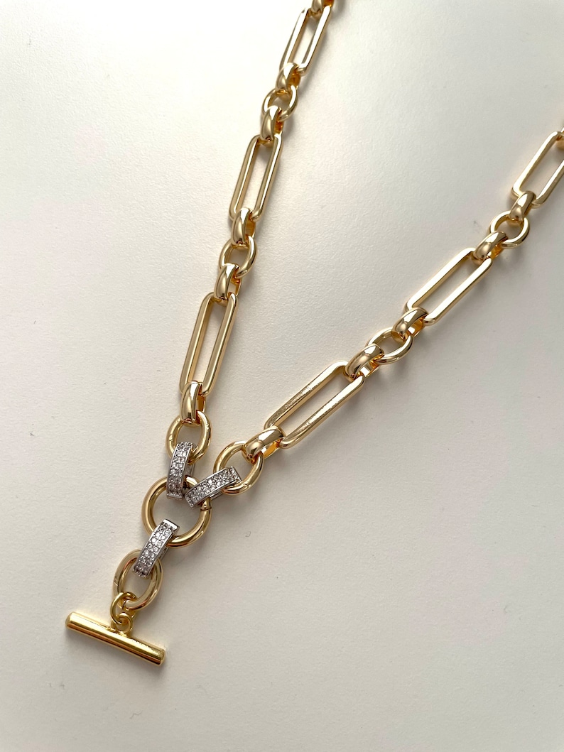 May include: A gold chain necklace with a T-bar clasp and a decorative link featuring small clear stones.