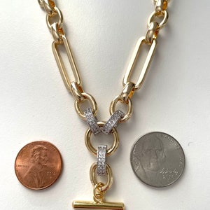 May include: A gold chain necklace with a large, rectangular link design. The necklace has a pendant with two rings that are set with clear cubic zirconia stones. The pendant has a small, gold bar attached to the bottom.