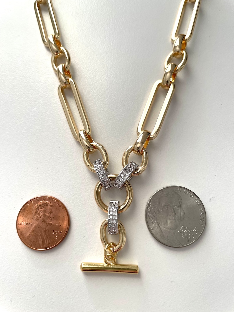 May include: A gold chain necklace with a large, rectangular link design. The necklace has a pendant with two rings that are set with clear cubic zirconia stones. The pendant has a small, gold bar attached to the bottom.